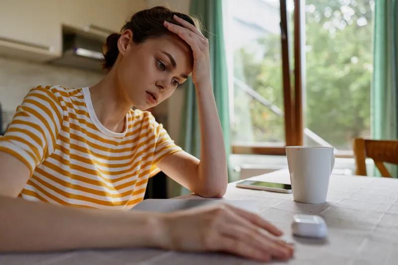 Stressed concentration a young woman in a striped shirt sits at a sunlit home table and gazes at a tablet, her hand on her forehead, conveying focus and fatigue in a cozy indoor space.