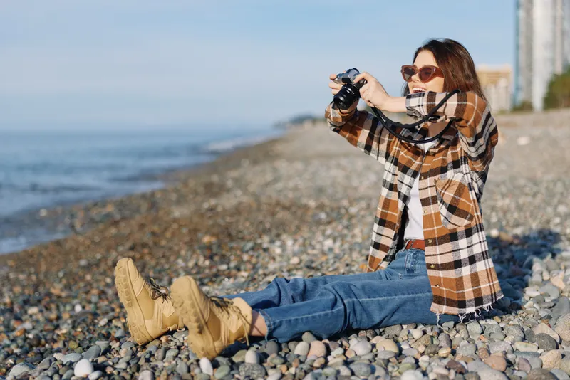 Photographer on a pebble beach captures a moment with a camera, wearing a plaid coat, jeans and boots, enjoying outdoor photography and relaxed leisure by the seashore.