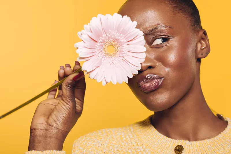 Close-up of a dark skinned African woman holding a pink flower near her face, wearing a yellow sweater. Bright yellow background highlights natural beauty and floral concept.