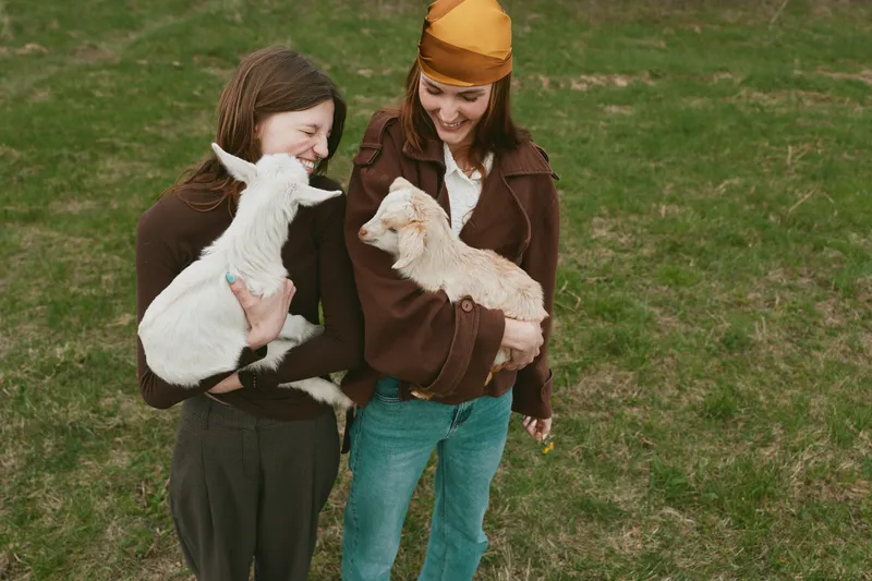 Two women in casual clothing hold fluffy goat kids in a sunny field, sharing smiles. A warm lifestyle moment capturing playful bonding with animals and nature, perfect for family, friendship, and