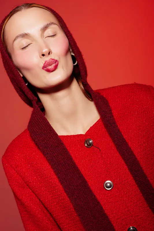 Young woman in red coat and scarf with closed eyes and red lips blowing a kiss on red background. Concept of fashion, beauty, and playful expression in studio lighting.