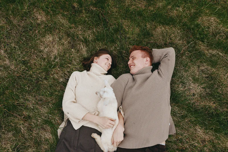 Lifestyle couple outdoors on grass with a small white goat kid, cozy sweaters, smiling faces, sunlit moment, relaxed posture, natural colors, lush background, gentle breeze, warm atmosphere