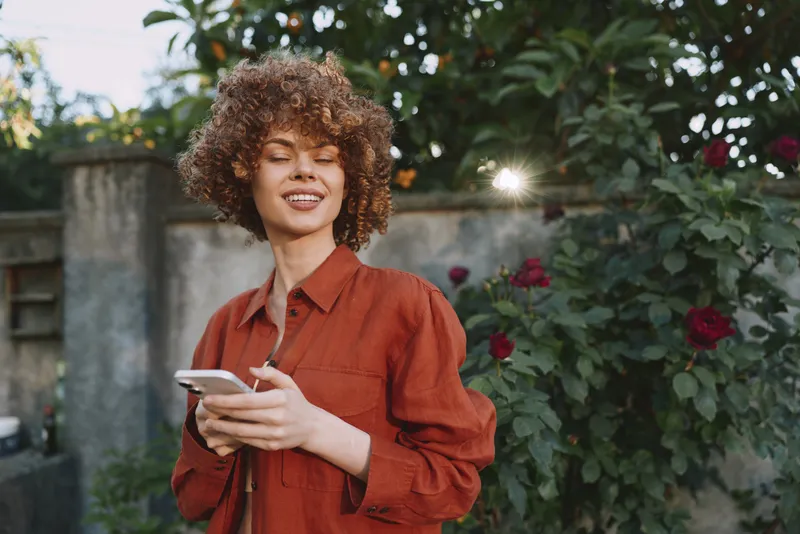 A young woman with curly hair in a rust shirt smiles while checking her phone in a sunny garden, conveying casual everyday connection and relaxed lifestyle