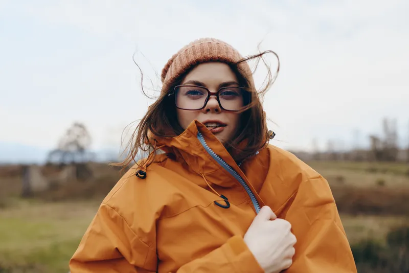 Woman lifestyle portrait outdoors in autumn countryside, orange jacket, beanie and glasses, holding zipper collar, windblown hair, cloudy sky and blurred field background