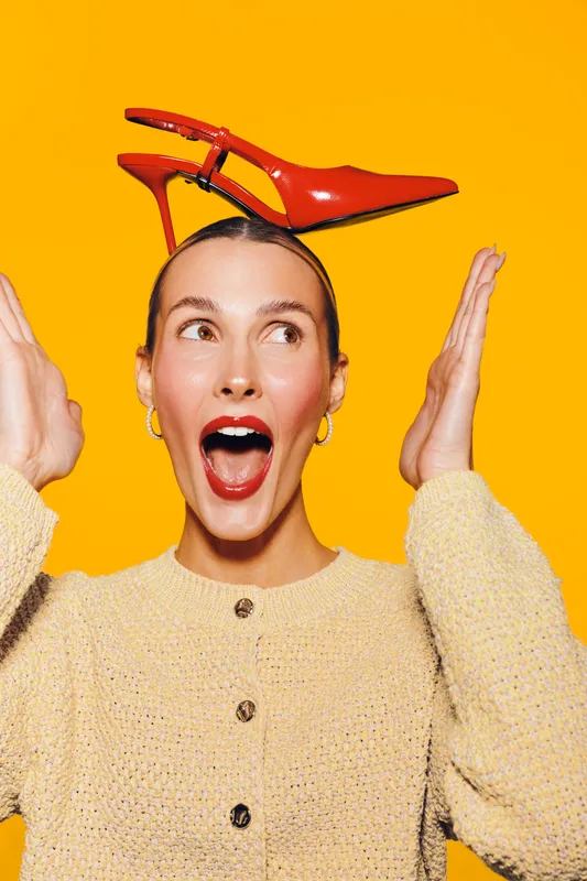Young woman with red high heel shoe on head looks surprised and excited. She wears yellow sweater and red lipstick on bright yellow background. Concept of fashion and fun.