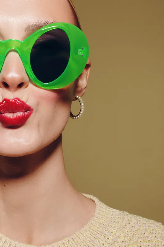 Close-up of a woman wearing bright green sunglasses and red lipstick making a kiss face. Beige background and textured sweater create a stylish and playful fashion portrait concept.