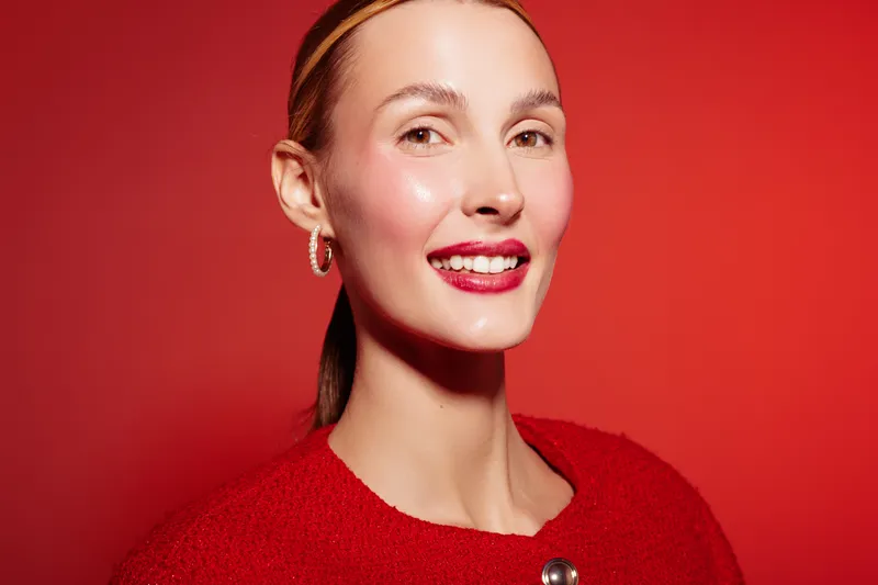 Smiling woman with red lipstick and earrings poses against red background wearing red sweater. Portrait of happy female model with natural makeup and shiny skin in studio lighting.