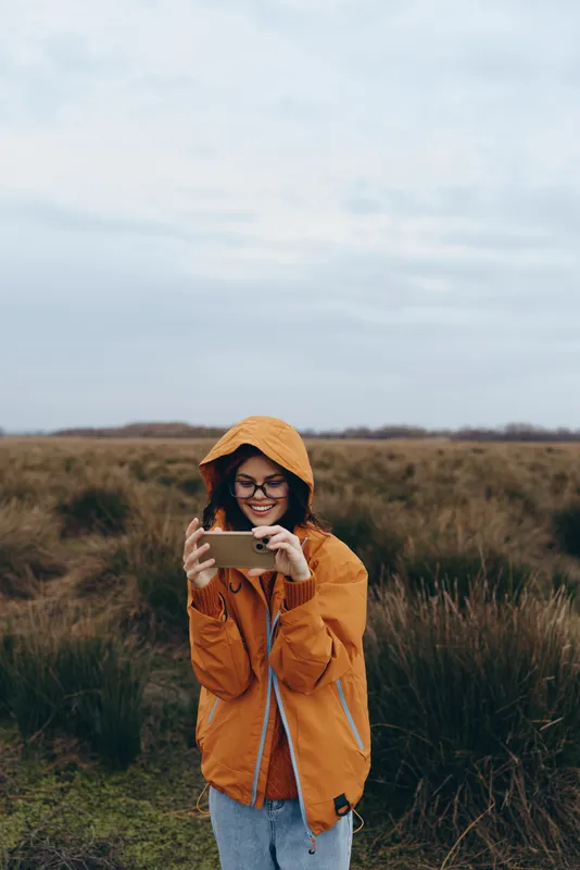 Woman in orange hood jacket and glasses with a smile holding a phone in a grassy field, outdoors lifestyle concept, natural light under cloudy sky, relaxed travel mood on weekends