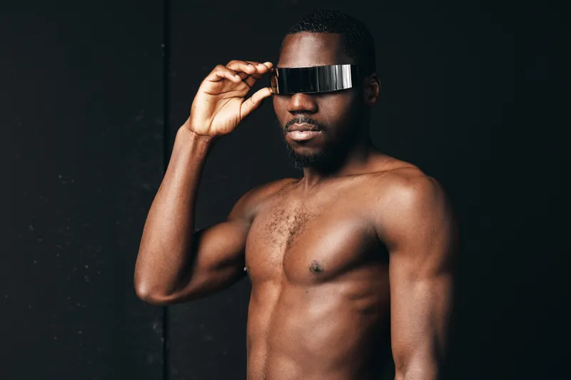 Creative conceptual portrait of shirtless African man wearing futuristic visor glasses in visual style with dark background and confident pose for modern fashion.