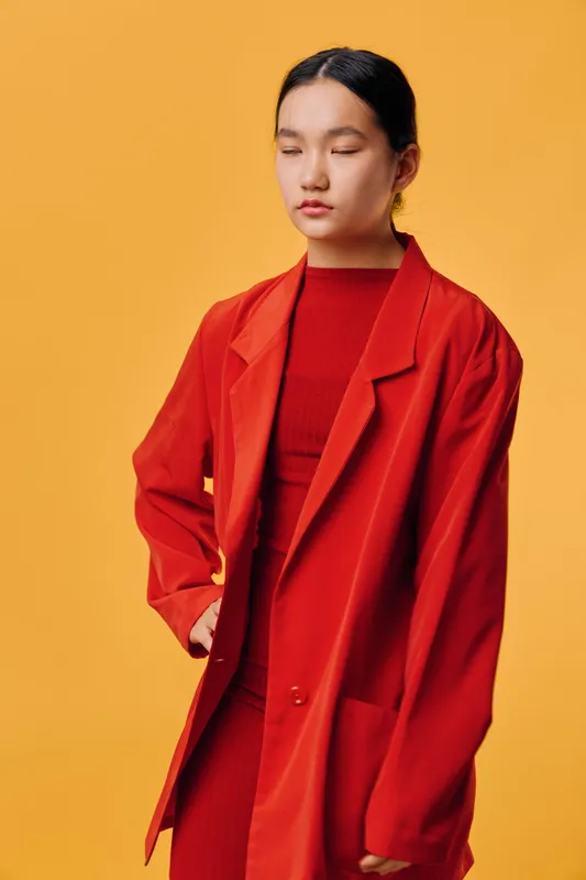 Young woman standing with eyes closed wearing red coat and matching dress against orange background, portrait of confident female model in casual fashion outfit for style concept.