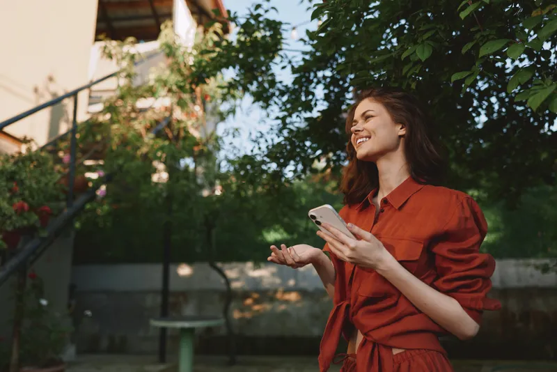 Cheerful woman outdoors, smiling in a red dress, holding a smartphone, sunlit moment with lush greenery, relaxed posture, soft breeze, natural colors, modern lifestyle vibe