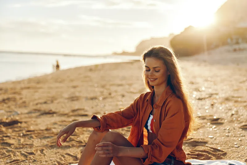 Young woman with long brown hair sits on a sunlit beach at sunset, wearing an orange jacket over a white top, relaxed and content as warm light enhances the mood.
