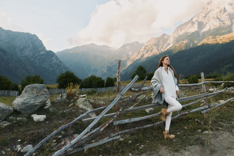 Young woman in white outfit and cozy jacket leans on rustic wooden fence while enjoying mountain landscape during early morning outdoors.