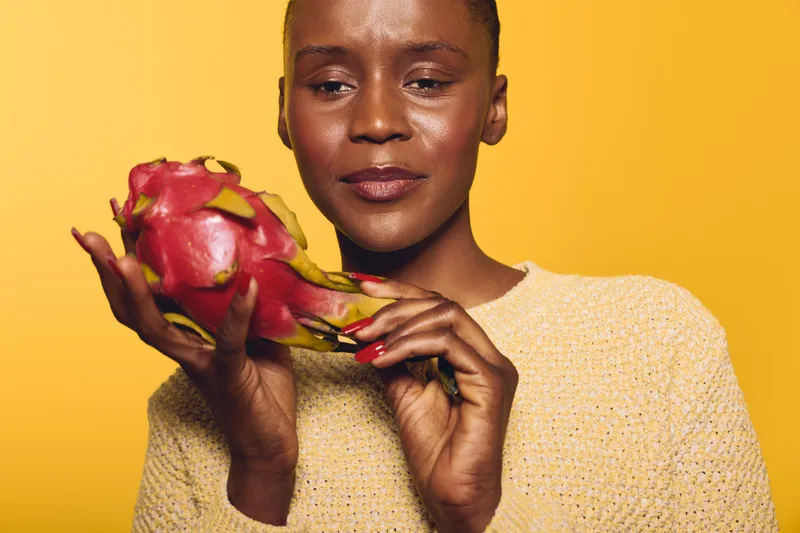 Close-up of a dark skinned African woman holding a red dragon fruit against yellow background. Concept of healthy lifestyle, exotic fruit, and natural beauty with vibrant colors.