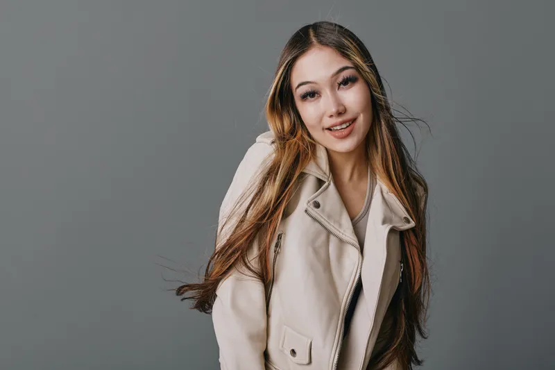 Portrait of confident young woman wearing light jacket smiling with long hair against gray background in studio. Happy casual female model posing for camera indoors.