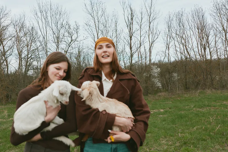 Goat, kid, outdoors, two women hold playful goats in a grassy field, smiling warmly, illustrating a lifestyle of friendship and rural connection in an open meadow