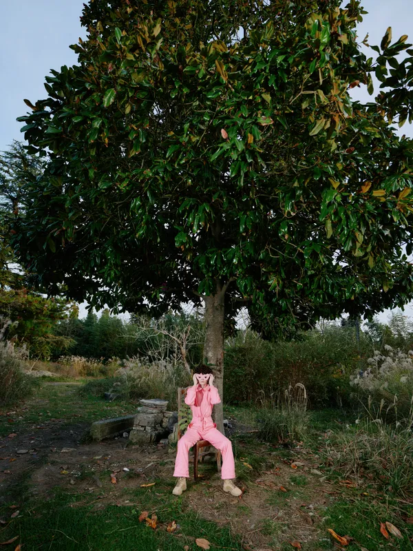 Woman in pink outdoor attire sits under a tree in a peaceful park, posing playfully, natural light and green foliage create a calm landscape, conveying relaxed leisure and outdoor joy
