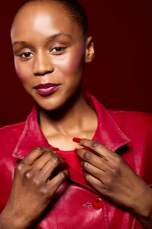 Close-up of a confident black woman with dark skin wearing a red leather jacket and red nail polish. She poses against a deep red background, showcasing fashion and beauty concepts.