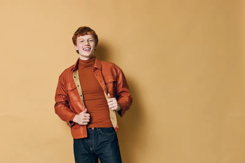 Young man in casual outfit with red hair smiling and standing against beige background, wearing orange jacket and turtleneck for fashion portrait.