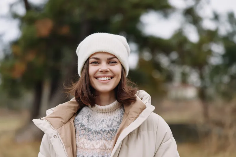 Woman smiling outdoors in a beige jacket and white beanie, portrait in a park during cool weather, casual style and cheerful mood.