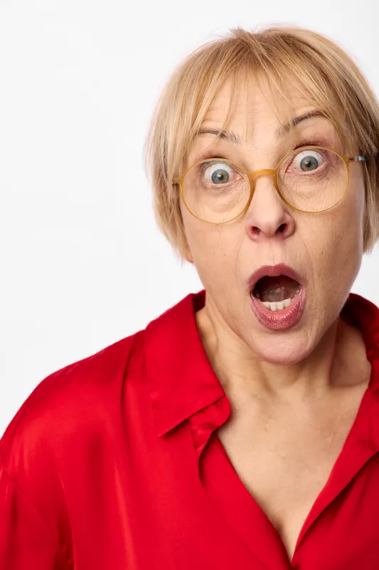 Surprise shocked woman with blonde senior look, round glasses, open mouth and wide eyes, red shirt, close up portrait, isolated on white background