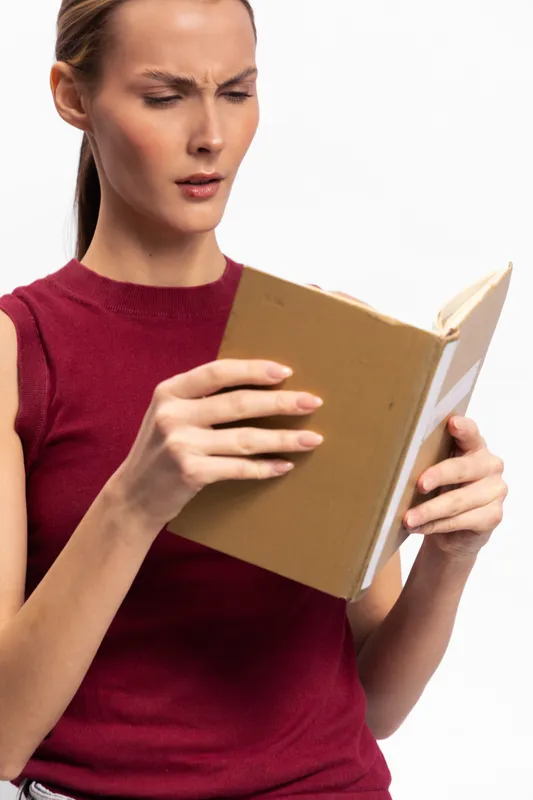 Young woman in red sleeveless top looks confused while reading a book. Isolated on white background. Concept for concentration, study, curiosity, and problem solving.