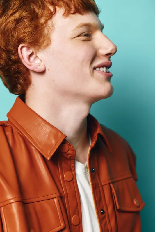 Redhead man with curly hair wearing brown leather jacket and white shirt smiling sideways on turquoise background. Casual style, portrait, youth fashion and natural expression in studio.