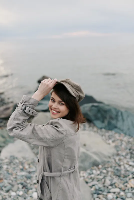 Smiling woman in beige coat and cap looks back at camera on rocky beach. Overcast sky and calm sea create serene mood. Concept for outdoor fashion, autumn, and lifestyle.