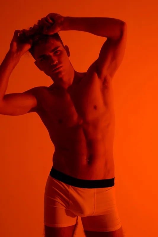 Young man with muscular body posing shirtless in white underwear against orange background. Fitness, male model, toned physique and confident expression in studio lighting.