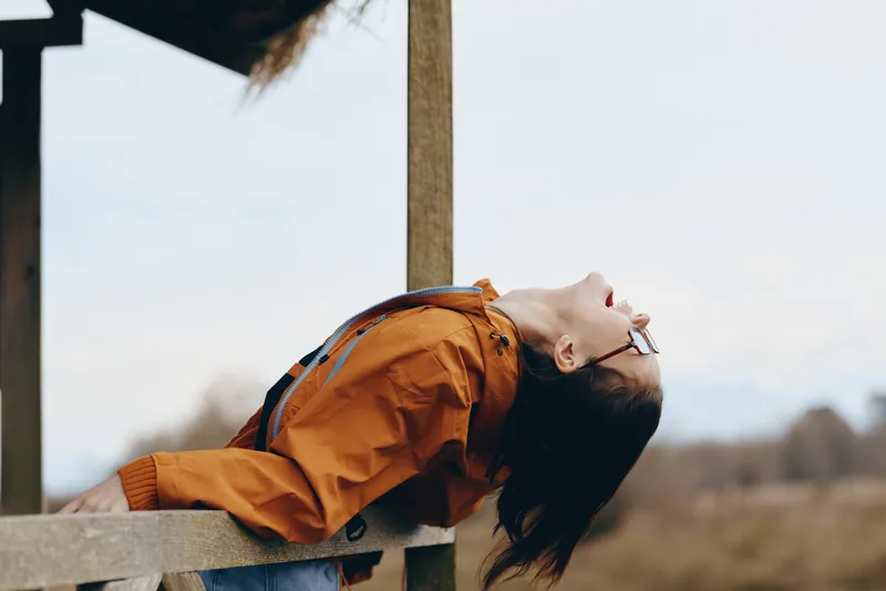 Woman lifestyle outdoors, leaning on wooden fence, wearing orange jacket, soft wind in hair, looking upward with calm smile, daylight portrait, natural background with sky copy space