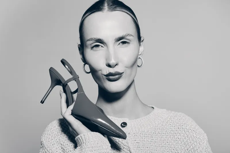 White woman with fair skin and slicked hair holds high heel shoe near face with confident expression on light background. Concept for fashion, style, and footwear accessories.