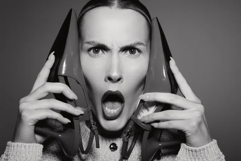 Young white woman with light skin holds two shoes near face with open mouth and intense expression.
