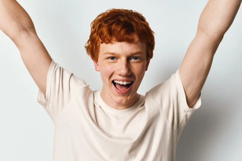 Young man with red hair wearing white t shirt raising arms and smiling broadly against white background for celebration and happiness concept.