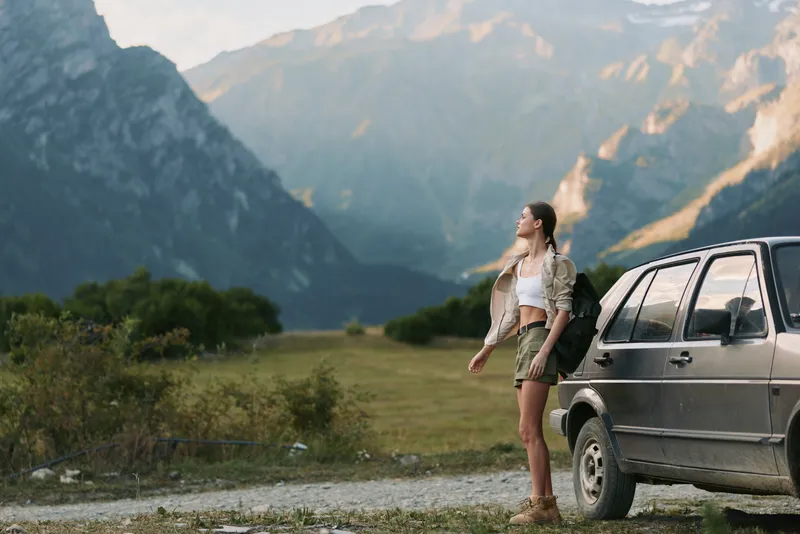 A woman in casual outfit stands beside a car on a dirt road looking at mountain scenery. The photo captures outdoor lifestyle and adventure in nature.