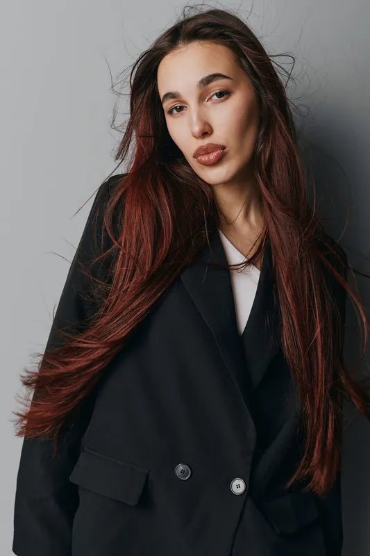 Young woman with long red hair wearing black blazer and white shirt posing against gray background in studio. Fashion model with natural makeup and confident expression looking at camera.