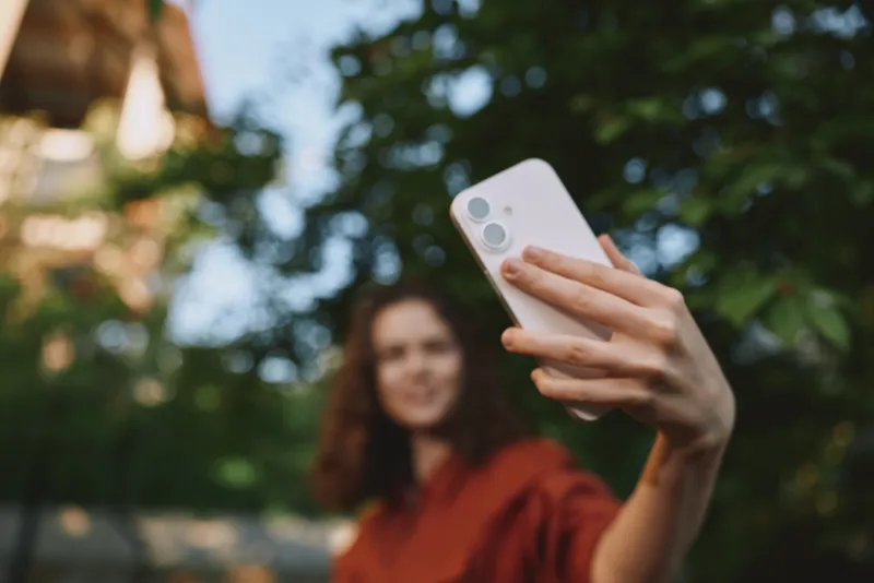 Woman takes a candid selfie outdoors in a park, smiling into her phone, capturing a casual lifestyle moment in natural light against a lush green backdrop