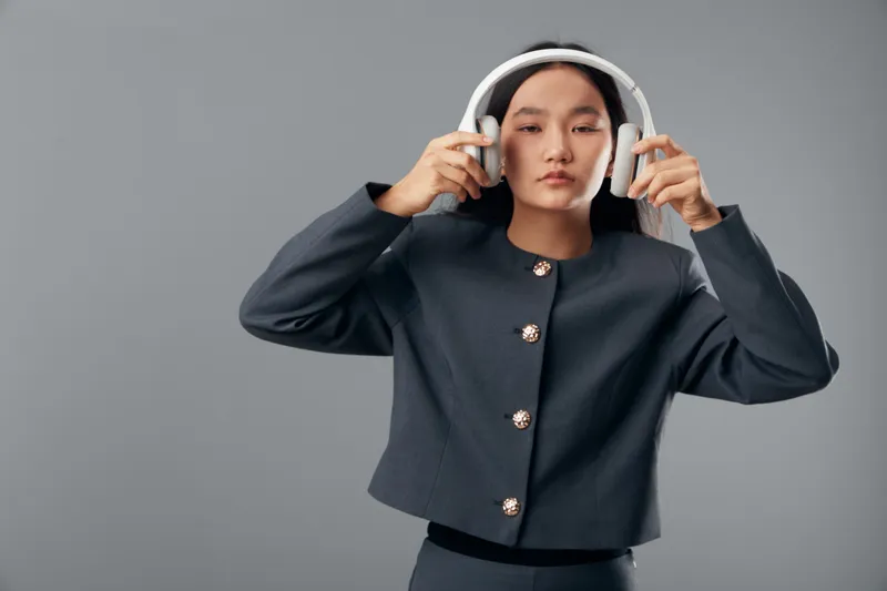 Young woman wearing formal dark jacket holds white wireless headphones near ears with eyes closed standing against gray background. Clear style technology and music concept.