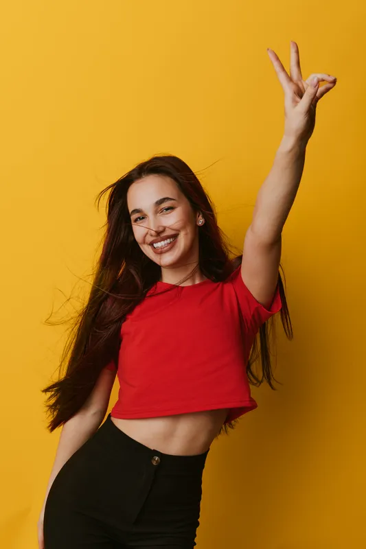 Young woman with long hair smiling and posing with raised hand on yellow background. Casual clothing, happy expression, and confident gesture in studio portrait photography.