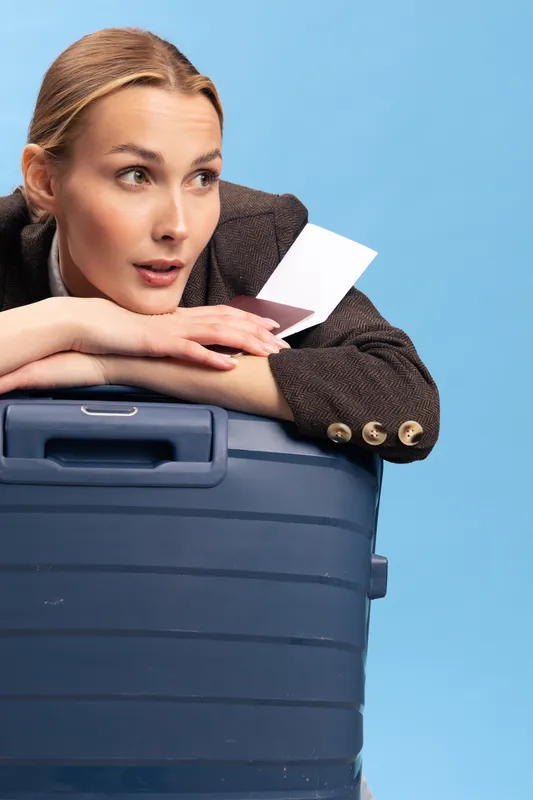 Young woman in brown jacket leans on blue suitcase holding passport and ticket, looking away. Concept for travel, vacation, planning, and anticipation of journey.