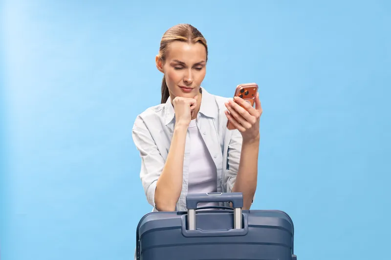 Young woman with suitcase looks at smartphone screen while waiting, isolated on blue background. Concept of travel planning, trip preparation, and modern communication technology.