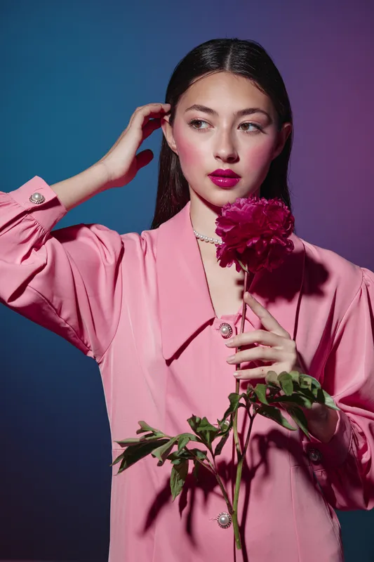 Young woman in a pink blouse holds a large red flower, styled with vibrant makeup against a gradient blue-purple background. Concept of creative beauty and fashion.
