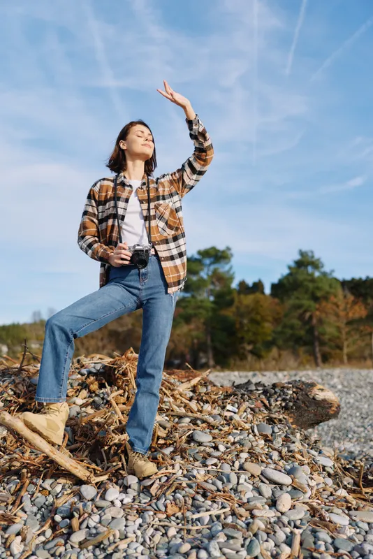 Woman outdoor camera nature casual plaid shirt jeans pose on rocky shore under bright sky, capturing a moment of exploration and freedom in open air