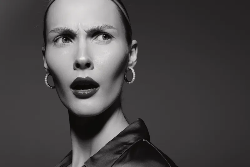 Confused young white woman with pale skin looks aside with raised eyebrows and open lips in black and white portrait.