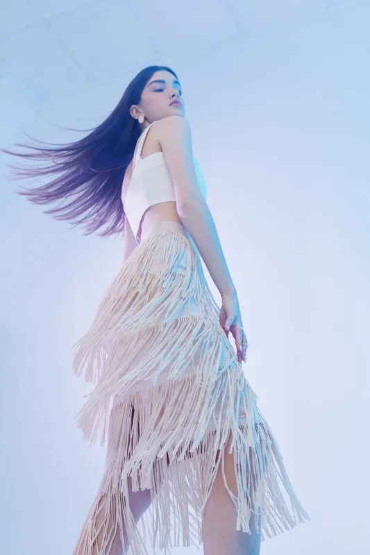Young woman beauty fashion model poses in a fringed skirt and white top. Her hair flows as she looks away in a soft studio with pastel lighting. Concept for style and elegance.