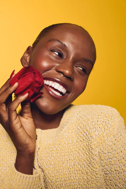 Happy African woman with dark skin holding red rose near face smiles with closed eyes on yellow background. Concept of beauty, joy, skincare, and natural happiness in studio portrait.