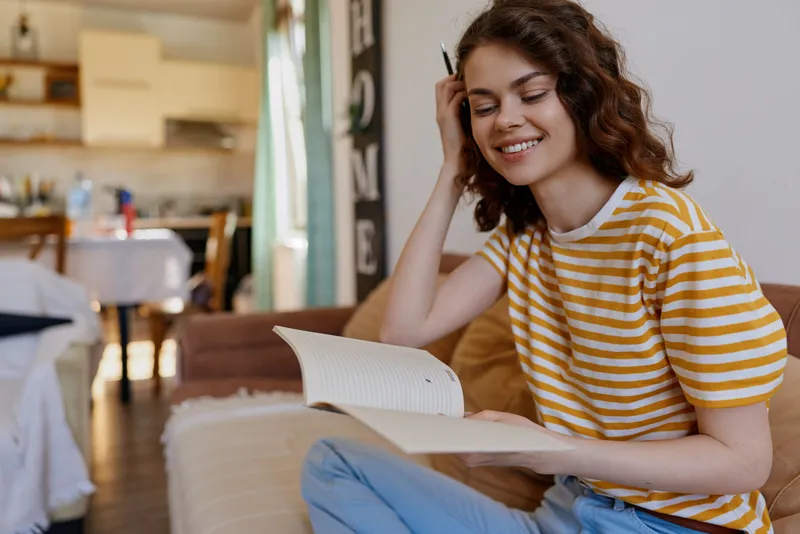 Young woman with curly hair smiles while reading a notebook on a cozy sofa at home, wearing a striped yellow tshirt, as natural light from a sunny window creates a relaxed and focused mood.