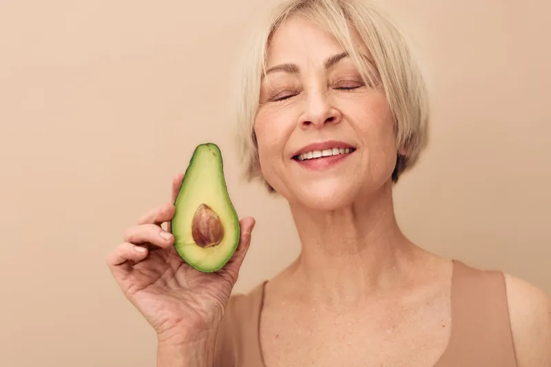 Smiling mature woman with short blonde hair holds fresh avocado half near face with closed eyes. Concept for healthy lifestyle, natural beauty, wellness, nutrition, and organic skincare.
