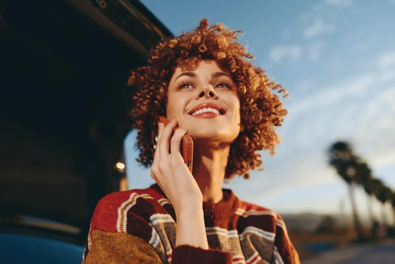 Woman lifestyle curly hair phone call smile outdoors street sunlight, relaxed portrait smiling young woman holds smartphone to ear, warm daylight city background, casual cozy jacket