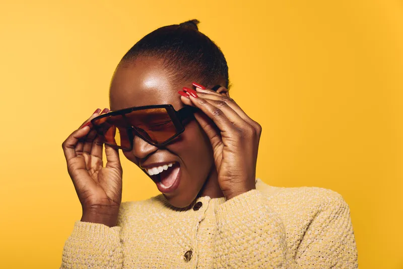 Happy African woman with dark skin laughing and adjusting large sunglasses. She wears a yellow sweater and stands against a bright yellow background. Concept of joy and fashion.
