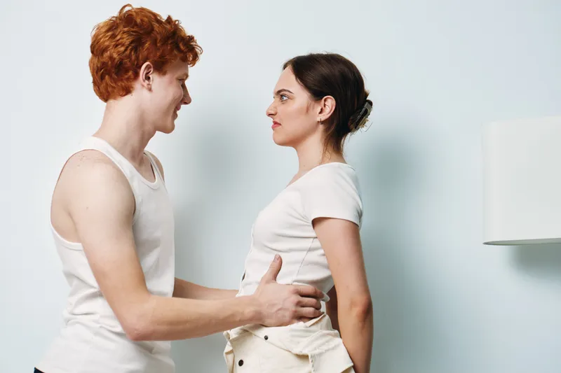 Couple standing face to face indoors with gentle touch on waist, young man and woman wearing casual clothes, interaction and connection in bright room with lamp.